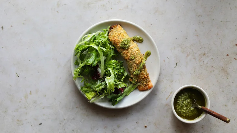 A piece of pesto-crusted baked salmon on a plate with a side salad and a drizzle of pesto next to a bowl of pesto