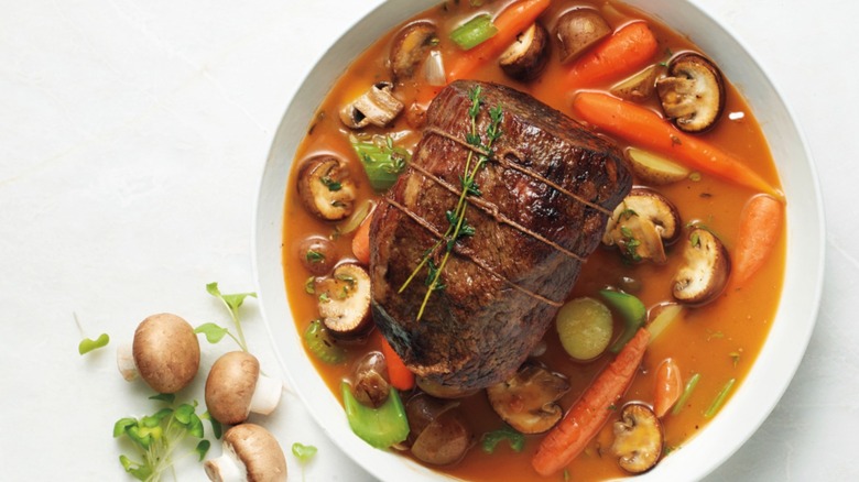 White dish holding pot roast with carrots, mushroom, and celery in broth on a white background with raw mushrooms and sprouts