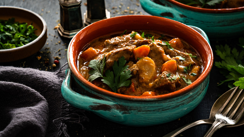 a bowl of beef and vegetables in rich gravy topped with fresh herbs and surrounded by cutlery, napkins, and other bowls
