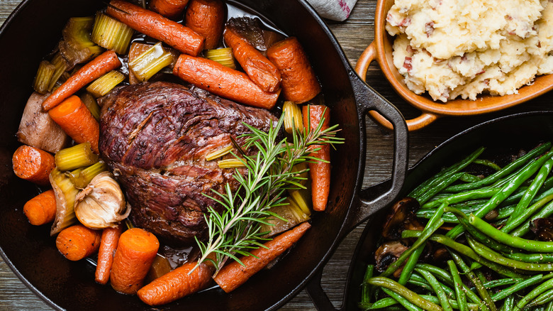 pot roast with vegetables in a cast iron, with dishes of mashed potatoes and green beans beside it
