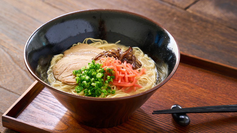 a bowl of ramen with pale broth and lots of toppings on a wooden tray with chopsticks