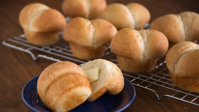 A sliced, muffin-shaped Rhodes dinner roll topped with melting butter sits on a plate in front of a rack of rolls.