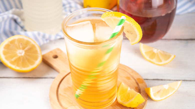 A yellow cocktail in a glass, with a green and white straw, garnished with a lemon wedge, sitting on a wooden trivet