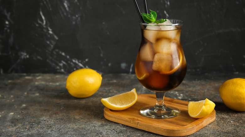 A cocktail glass full of a dark brown liquid and ice cubes, garnished with mint. There are lemon wedges sitting next to it on a wooden trivet and whole lemons on the table around it