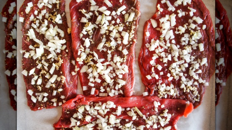 Raw steak is marinating in diced onions on a sheet tray.