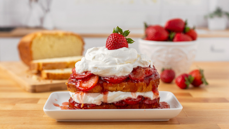 Saucy strawberries and whipped cream layered between sliced pound cake on a plate, in front of ingredients
