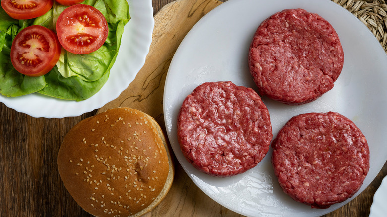 Raw burgers patties on plate, next to burger bun, lettuce, and sliced tomatoes