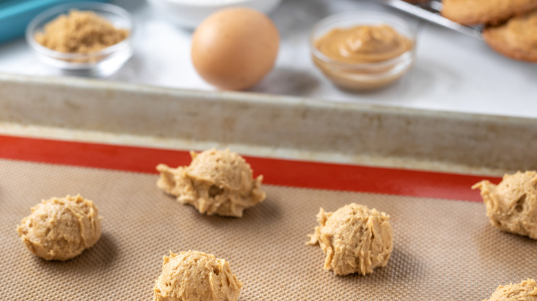 Balls of uncooked cookie dough sit before a bowl of brown sugar, a bowl of peanut butter, and a large egg
