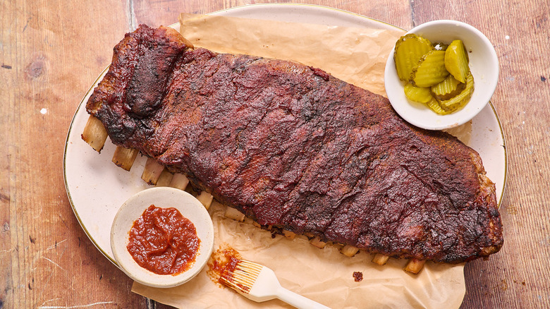 Aerial view of uncut St. Louis-style ribs on a platter with dishes of pickles and sauces on the side