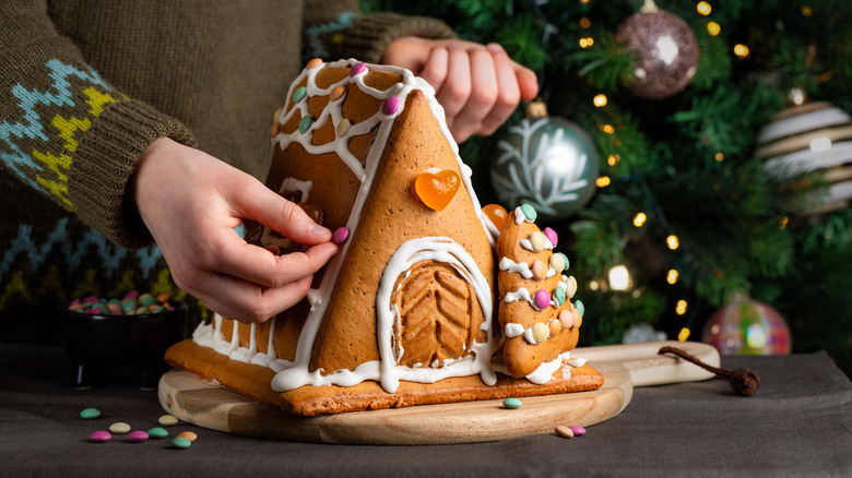 A person in a festive holiday sweater decorating a small gingerbread house
