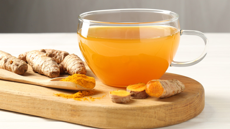 A glass cup of turmeric tea with ground and fresh turmeric on a chopping board