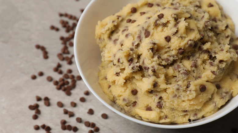 Chocolate chip cookie dough in bowl with chocolate chips on the table in the background.