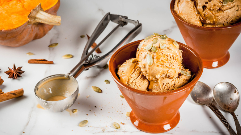 Two brown sundae bowls of pumpkin ice cream next to a scoop and leftover toppings