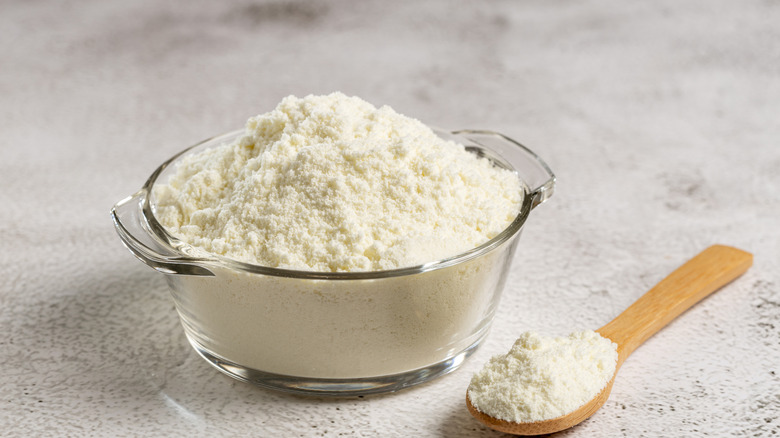 Milk powder in a glass bowl with a wooden spoon of it nearby.