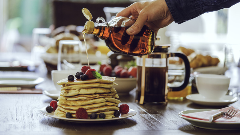 Person pouring maple syrup from glass bottle onto stack of pancakes with fersh berries