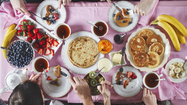 Overhead shot of people eating breakfast at a table with lots of pancakes, fruits, crepes, jams, and syrup