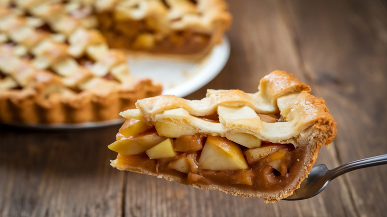 Slice of apple pie with latticed crust balanced on a fork, with the rest of the pie blurred in the background