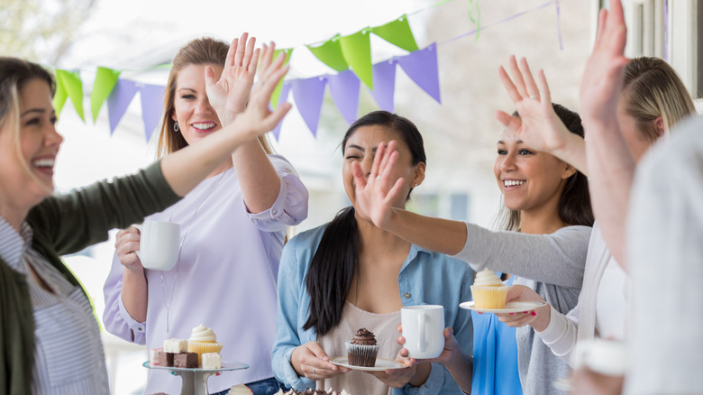 Group of women high-fiving over trays of dessert and holding cups of coffee under a banner