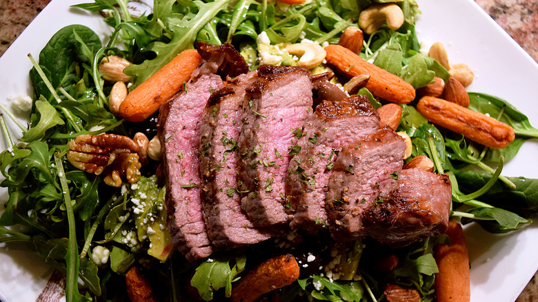 A plated London broil, with the steak sliced and arranged on a bed of salad, carrots, and nuts