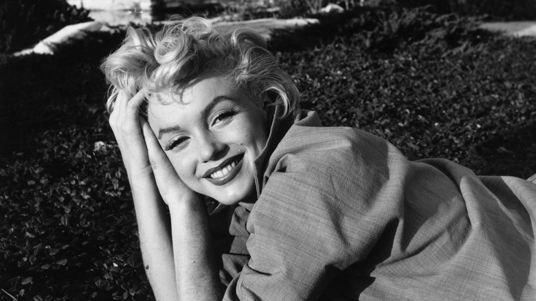 Marilyn Monroe lies in the grass with her hands clasped beside her head, smiling at the camera