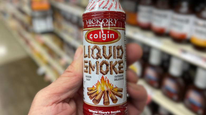 A hand holds a bottle of hickory-flavored Colgin Liquid Smoke in a grocery store aisle, with shelves in the background