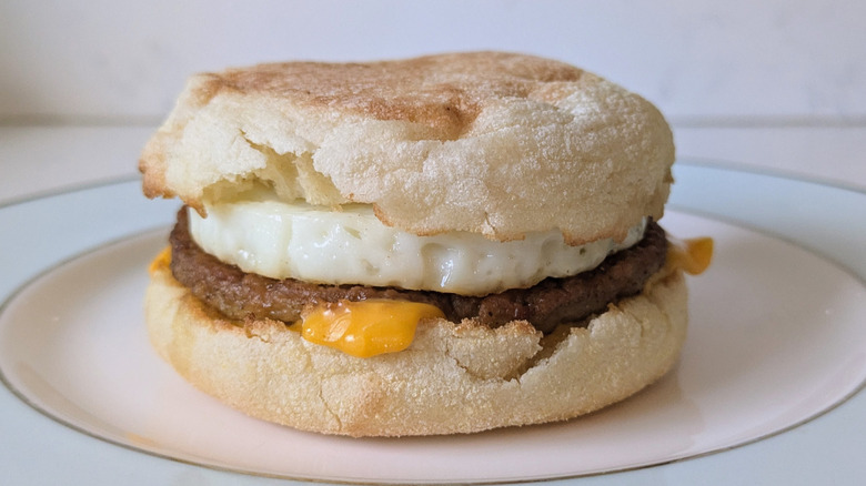 An English muffin is sliced and toasted, topped with a fried egg, a sausage patty, and a slice of cheese