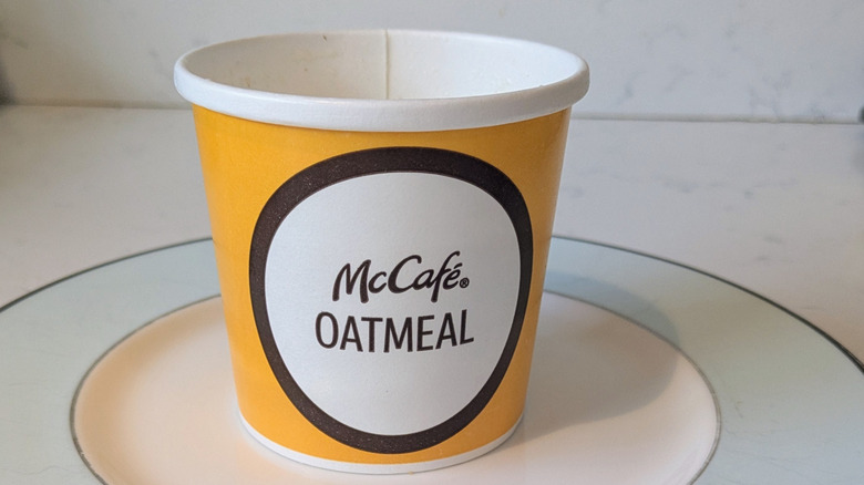 A McDonald's branded McCafe Oatmeal cup, yellow, white, and dark brown, sits on a blue and white plate. No oatmeal is visible  as the cup was not filled to the top