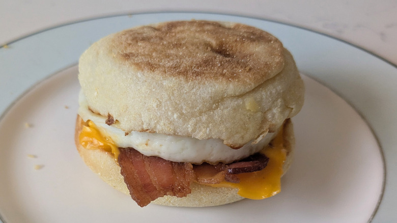 An English muffin is sliced in half, toasted, and topped with a slice of cheese, bacon, and a fried egg