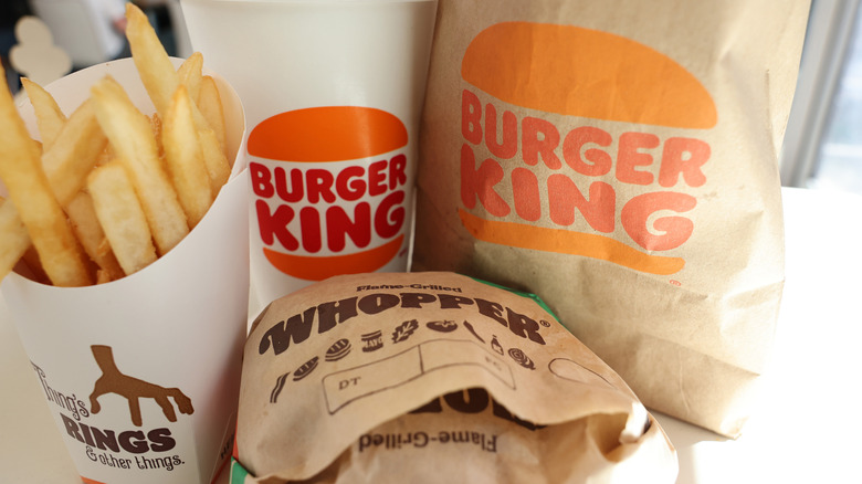 A Burger King Whopper wrapped in paper sitting in front of a Burger King drink, paper bag, and next to a carton of french fries