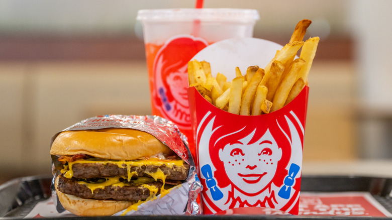 A Wendy's meal featuring a double cheeseburger wrapped in paper, a carton of french fries, and an orange drink in the background