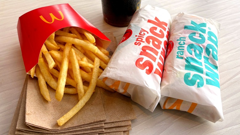Two wrapped McDonald's Snack Wraps alongside red sleeve of fries and stack of brown napkins on wooden table