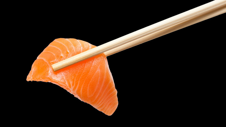 A piece of sashimi grade salmon being held between chopsticks against a black background