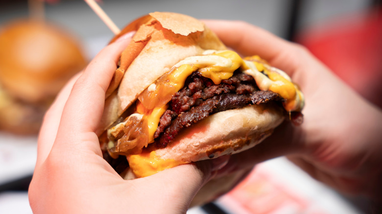 Two hands holding a large double cheeseburger wrapped in paper