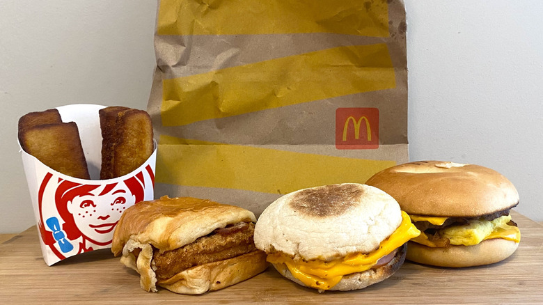 Wendy's Homestyle French Toast Sticks and Maple Bacon Chicken Croissant next to McDonald's Egg McMuffin and Steak, Egg and Cheese Bagel with a McDonald's bag in the background.