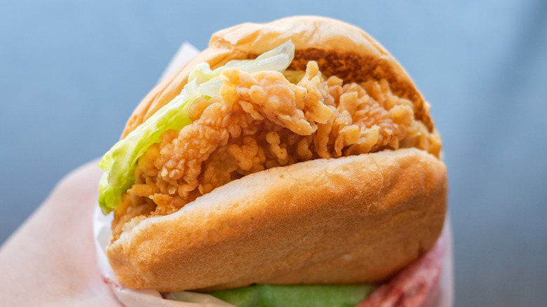 A close-up image of a hand holding a partially unwrapped fried chicken sandwich