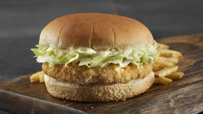 A fried chicken sandwich with lettuce and mayonnaise on a bun with fries behind it on a wooden cutting board