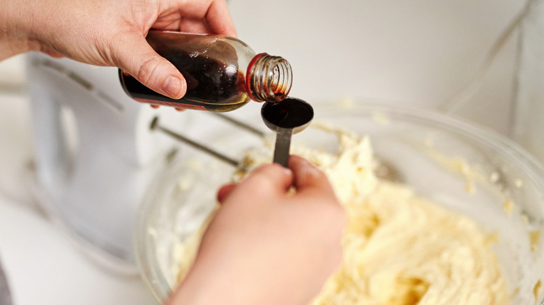 Someone pouring vanilla extract into a measuring spoon over a clear mixing bowl full of white batter