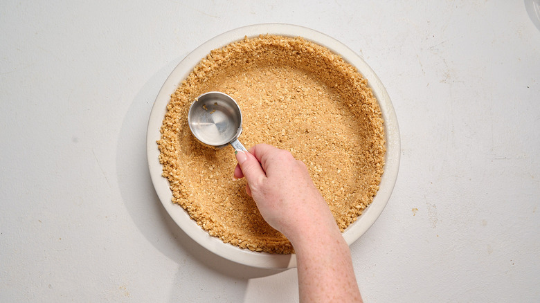 pressing crumbs into a pie pan with a measuring cup