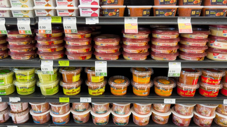 A refrigerated case of fresh salsas and hummus in plastic containers.