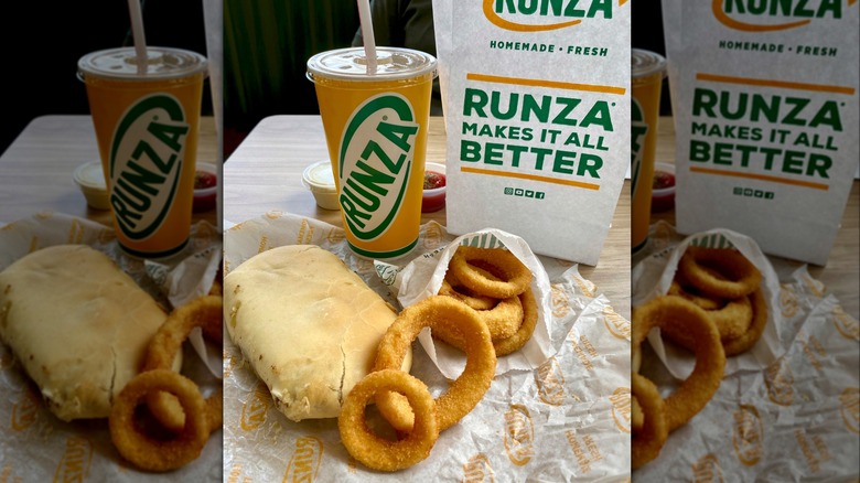 A Runza sandwich with some onion rings and a drink in front of a Runza paper bag sitting on a table.
