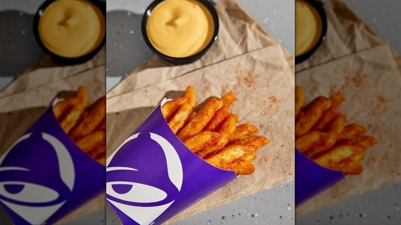 A purple cardboard box full of seasoned Taco Bell Nacho fries laying down on a napkin with a container of nacho cheese