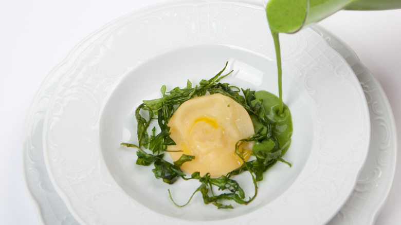 White plate on white table with piece of ravioli and green vegetables with green sauce