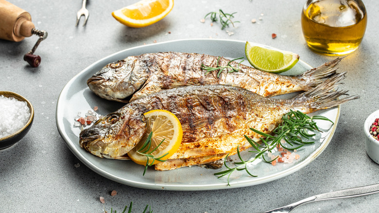Two whole grilled fishes on a white plate with lemon and herbs