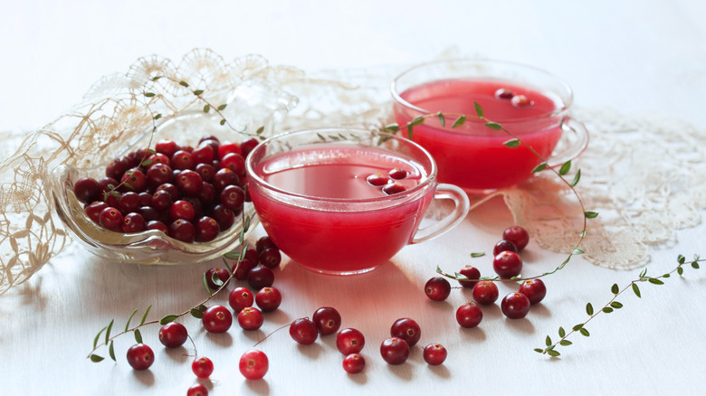 Cranberries in bowl and scattered on white table beside cranberry juice in teacups