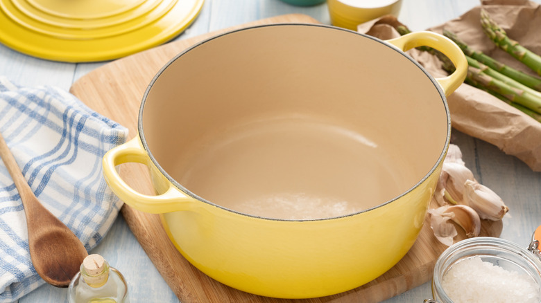 Yellow enameled Dutch oven with an off-white interior sitting on a cutting board, surrounded by ingredients and a wooden spoon