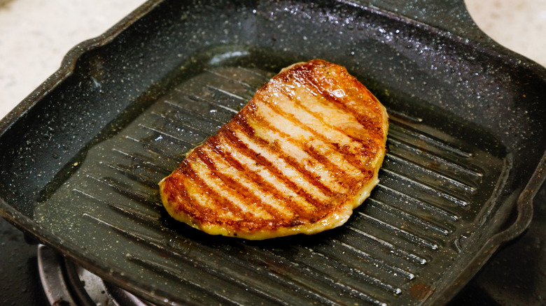 Pork chop or chicken breast cooking in a square, well-used, cast-iron grilling pan, with distinct, well-seared grill marks on the meat