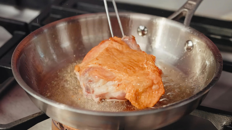 Chicken or turkey thigh being seared in shallow oil in a stainless-steel skillet over a gas burner