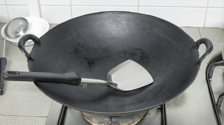 Empty but well-seasoned, carbon-steel wok sitting on a stovetop gas burner, with a wok spatula resting inside the pan