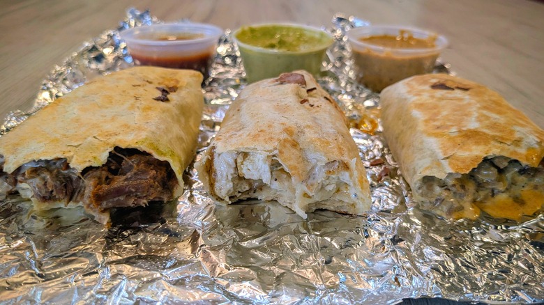 Three half-eaten mini burritos on a piece of tin foil with cups of sauces behind them