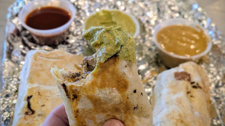 A hand holds a burrito with a bite taken out and green sauce on it with sauce cups in the background